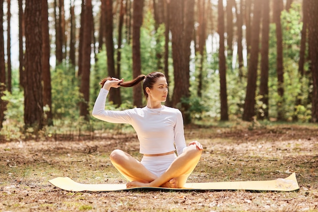 Attractive confident female wearing white top and leggins sitting on keremat on ground with crossed legs in lotus pose, looking thoughtfully aside, touching her ponytail.