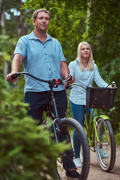 An attractive couple of a blonde female and man dressed in casual clothes on a bicycle ride in a park.