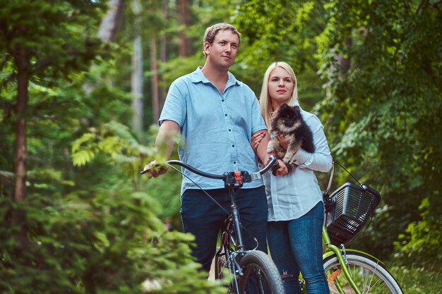 Привлекательная пара блондинки и мужчины, одетые в повседневную одежду, катаются на велосипеде со своим милым маленьким шпицем в парке.