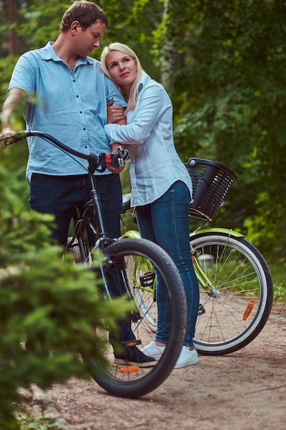 An attractive couple of a blonde female and man dressed in casual clothes, embrace near a bicycle in a park.