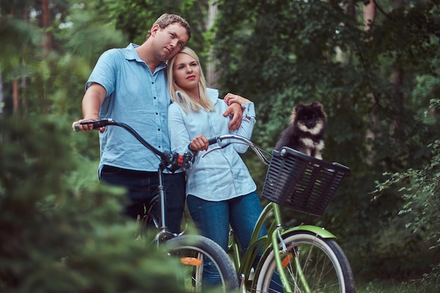 An attractive couple of a blonde female and man embrace, dressed in casual clothes on a bicycle ride with their cute little spitz in a basket.