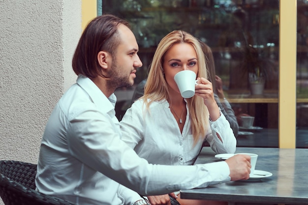 Attractive couple - charming blonde female dressed in a white blouse and bearded male with a stylish haircut dressed in a white shirt during a date at a cafe outdoors.