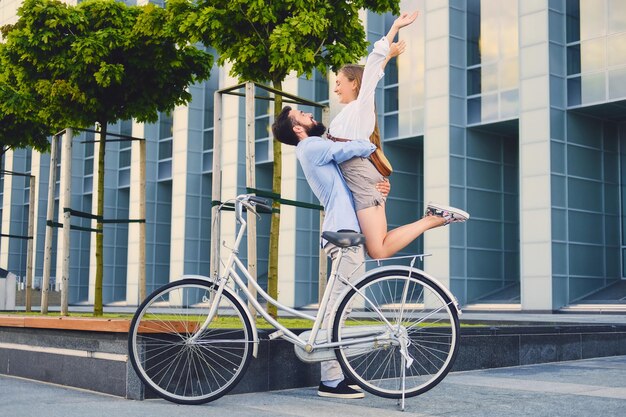 도시에서 자전거를 탄 후 데이트를 하는 매력적인 커플. 남자는 현대적인 건물 배경 위에 여자를 껴안습니다.