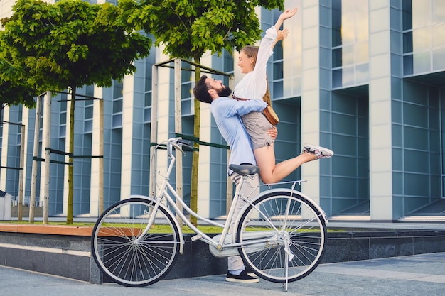 街で自転車に乗った後のデートで魅力的なカップル。男性は、モダンな建物の背景に女性を抱きしめます。