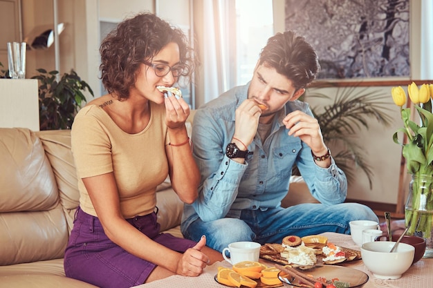 Attractive couple, handsome bearded stylish guy and curly beauty girl eating a meal at home while sitting on a sofa.