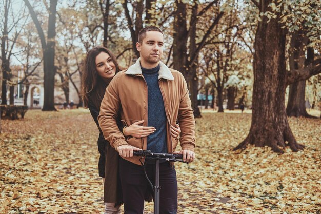 魅力的なカップルはスクーターで秋の公園で素敵なロマンチックな散歩をしています。