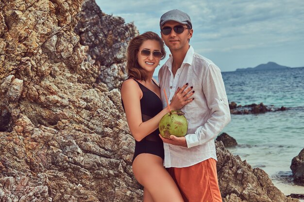 魅力的なカップルは、チューブでココナッツを持って、大きな岩礁の石の近くのビーチに立って、美しい島での休暇を楽しんでいます。