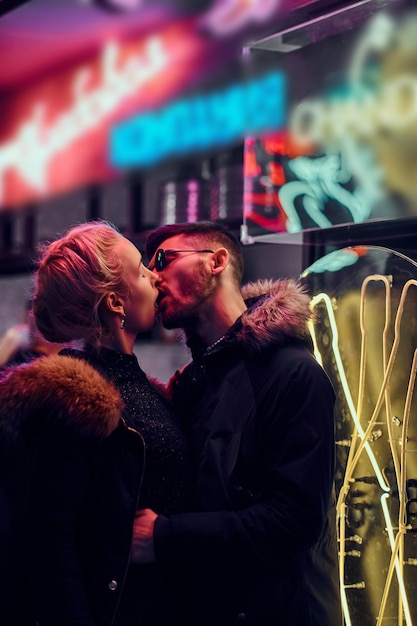 Attractive couple in love. A beautiful girl kisses her boyfriend standing in the night on the street. Illuminated signboards, neon, lights.