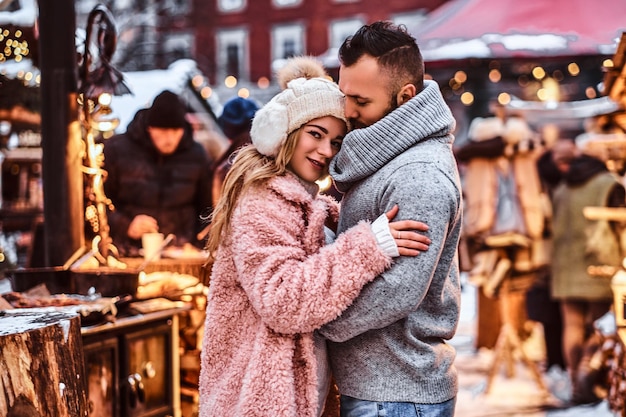 An attractive couple in love, a handsome man and charming girl cuddling and enjoying spending time together while standing at the winter fair at a Christmas time