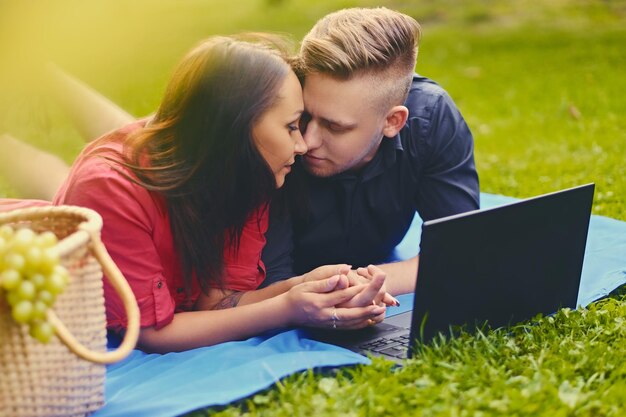 Привлекательная пара лежит на одеяле на лужайке и использует ноутбук на пикнике.