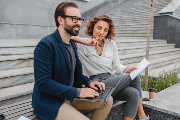 Attractive couple of man and woman sitting on stairs in urban city center, working together on laptop, smiling