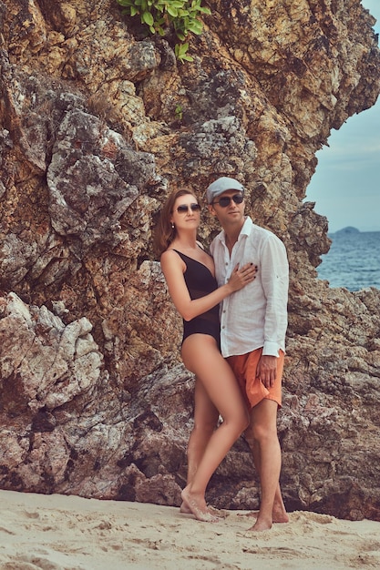 큰 암초 돌 근처 해변에서 포즈를 취한 매력적인 커플은 아름다운 섬에서 휴가를 즐깁니다.