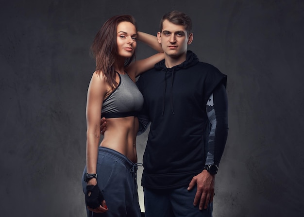 Attractive couple, slim brunette female and handsome guy in sportswear cuddling in a studio. Isolated on a dark background.