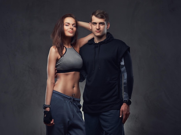 Attractive couple, slim brunette female and handsome guy in sportswear cuddling in a studio. Isolated on a dark background.