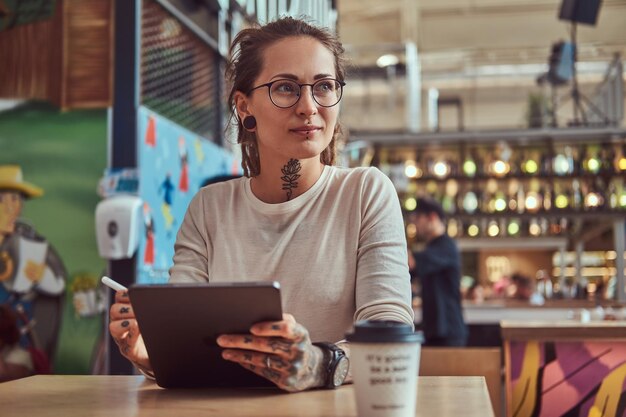 彼女の手に入れ墨を持つ魅力的な創造的な女の子は、彼女のデジタルメモ帳でスケッチしながらカフェに座っています。