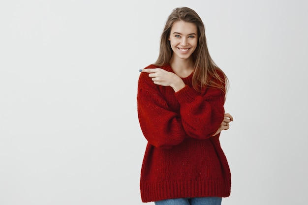 Attractive creative girlfriend has interesting idea. Pleased successful romantic woman in trendy loose sweater, pointing left with index finger and smiling broadly, standing confident over gray wall