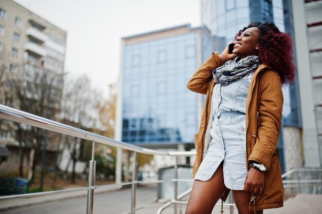 갈색 코트를 입은 매력적인 곱슬머리 아프리카계 미국인 여성이 휴대전화로 말하는 현대적인 다층 건물에 대해 난간 근처에서 포즈를 취했습니다.