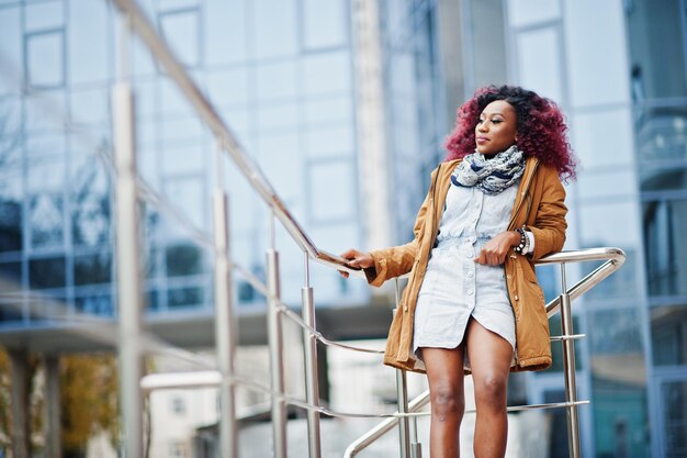 Attractive curly african american woman in brown coat posed near railings against modern multistory building