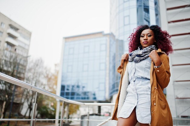 Attractive curly african american woman in brown coat posed near railings against modern multistory building