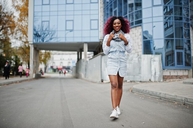Привлекательная кудрявая африканская американка в джинсовом платье позирует на фоне современного многоэтажного здания
