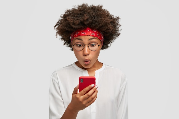 Attractive curly dark skinned female with afro hairstyle looks agitated at screen of smart phone, impressed by message content recieved from friend, has surprised facial expression, stands indoor