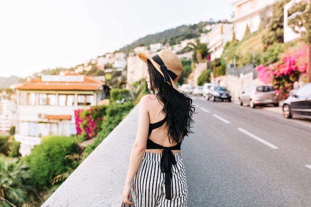 Привлекательная кудрявая девушка с длинными черными волосами, наслаждаясь видом на город, гуляет по дороге