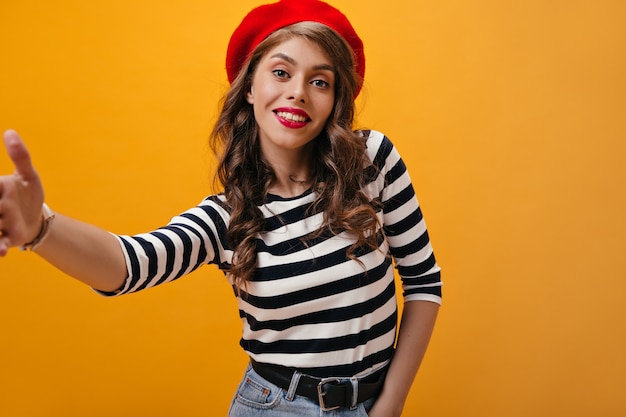 Attraente donna riccia in berretto fa selfie su sfondo arancione. ragazza moderna con capelli ondulati in maglione a righe in posa su sfondo isolato.