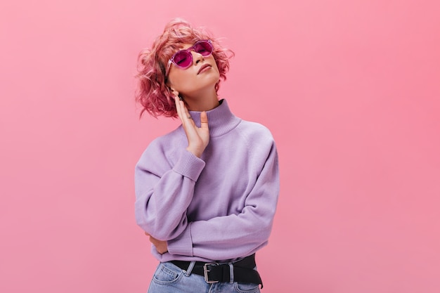 Attractive curly woman in purple cashmere sweater and fuchsia sunglasses poses on isolated wall