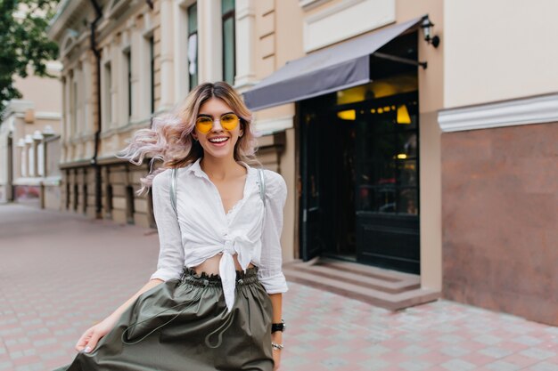 Привлекательная кудрявая женщина с искренней улыбкой играет со своей длинной юбкой во время прогулки по улице