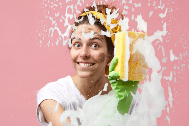 Attractive cute young Caucasian woman from cleaning service smiling broadly while tidying up in apartment, washing glass surface of window or shower stall, feeling excited and happy about her job