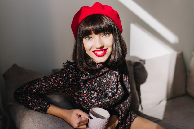 Attractive dark-haired girl in red beret and retro dress sitting in cozy room while drinking tea. Portrait of adorable brunette young woman in french outfit, gladly posing during coffee break at home