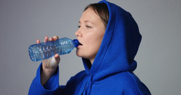 白い背景の上のプラスチック製の透明な青いボトルから水を飲む明るい青の特大パーカーの魅力的な黒髪の女性