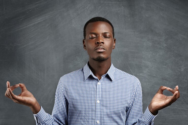 Free Photo attractive dark-skinned businessman with peaceful expression meditating, holding his hands in mudra gesture, keeping his eyes closed while practicing yoga