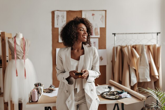 Attractive dark-skinned curly brunette woman in oversized jacket and white pants smiles, holds phone and leans on table in office of fashion designer