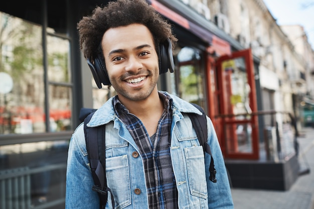Attractive dark-skinned smiling guy with bristle, listening to music while walking on street, being in good mood