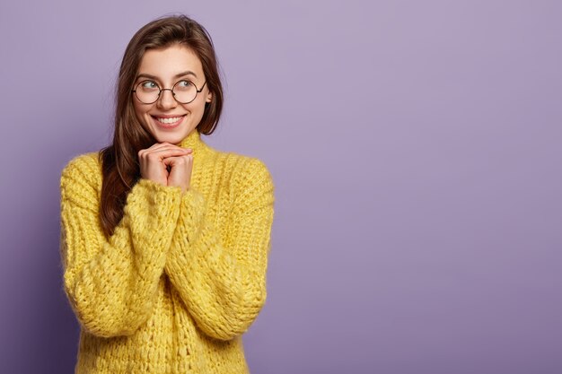 Attractive dreamy satisfied female holds both hands under chin, looks aside, feels glad