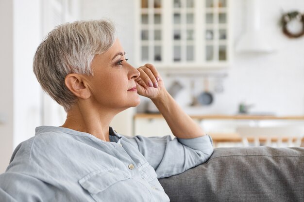 Attractive elegant gray haired female pensioner in stylish blue shirt sitting on sofa in living room, touching her face, thinking about her life. People, lifestyle, interior and coziness concept