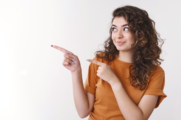 Attractive enthusiastic armenian young woman curly hair pointing looking intrigued left show index fingers awesome blank space advertisement smirking joyfully introducing product, white background