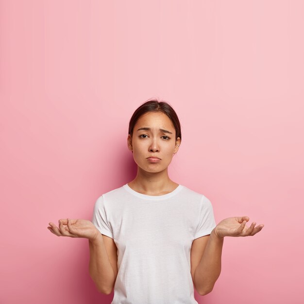 Attractive ethnic woman spreads palms with uncertainty, has unhappy look, faces difficult situation, dressed in white casual wear, poses against pink wall. People, doubt and unawareness