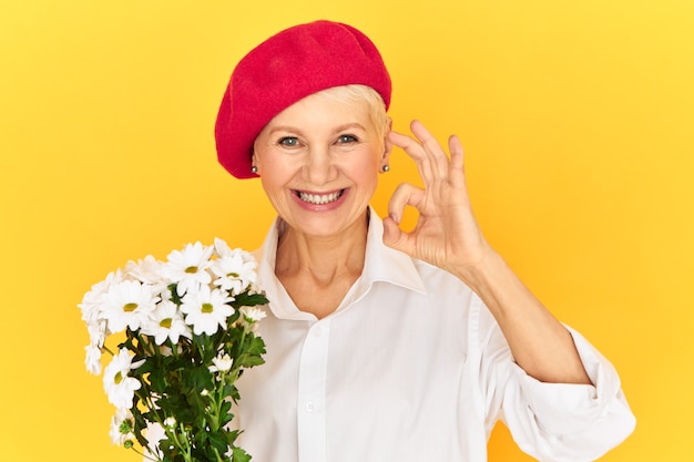 Attractive European female pensioner with blue eyes posing isolated in red headwear expressing delight, holding white daisies and showing ok gesture as sign of approval, smiling broadly at camera