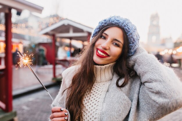 Attractive european lady in gray coat celebrating new year on the street, holding Bengal light.  Outdoor portrait of happy brunette girl with red lips posing with sparkler in winter.