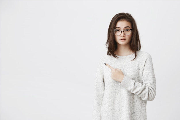 attractive european model pointing index finger aside with half-opened mouth, expressing curiosity and wonder, wearing glasses and trendy shirt..