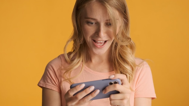 Attractive excited blond girl emotionally playing in game on smartphone over yellow background