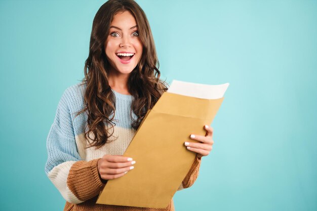 Attractive excited girl in cozy sweater  opening response envelope joyfully looking in camera over blue background