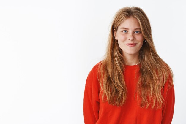 Attractive fair-haired girl with charming freckles and deep blue eyes smiling friendly, optimistic standing in warm cozy red sweater upright being in good mood listening, against grey wall.