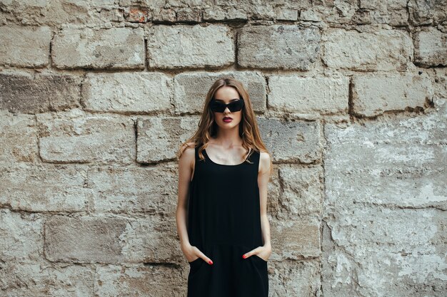 Привлекательная модная женщина в черном платье с солнцезащитными очками позирует возле белой стены