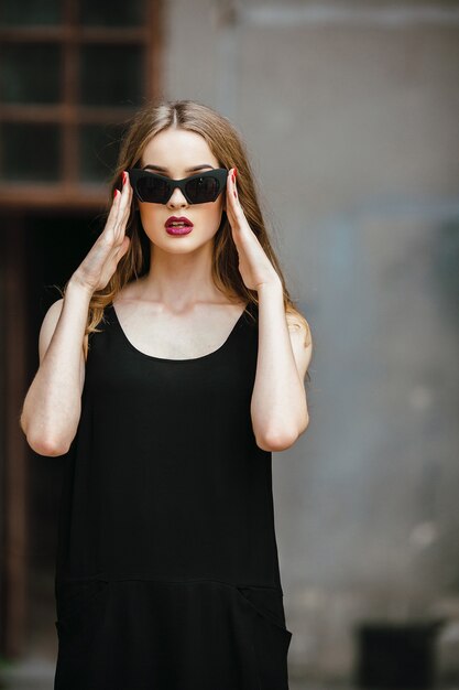 Attractive fashion woman in black dress with sunglasses posing near white wall