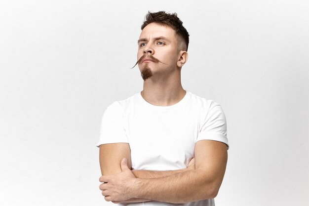 Attractive fashionable young bearded European hipster in white t-shirt crossing arms and looking up, being offended or insulted, say no words, waiting for your apologies. Portrait of proud haughty man