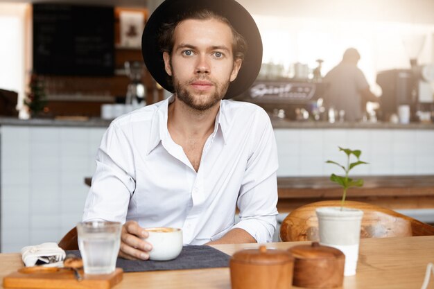 Attractive fashionable young man with beard having coffee at modern cafe