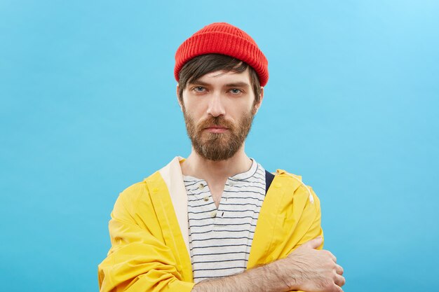привлекательный модный молодой человек с бородой, одетый в модную красную шляпу и желтый плащ, скрестив руки и имеющий серьезный уверенный вид. Люди, стиль и мода